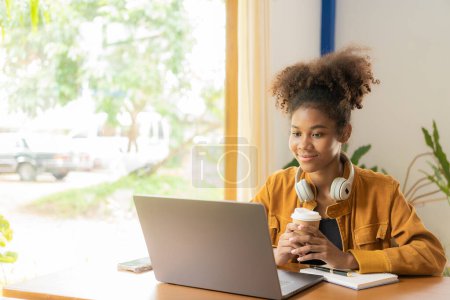 Téléchargez les photos : Jeune femme africaine étudiant en ligne avec ordinateur portable dans un café Millennial African American girl avec casque travaillant sur ordinateur portable dans un café - en image libre de droit