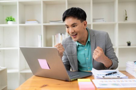 Photo for Happy young Asian businessman celebrates work success in office looking at laptop, excited about growth of financial trade. - Royalty Free Image
