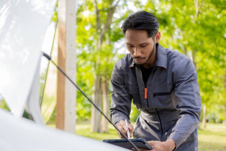 Photo for Auto mechanic inspects car by inspecting and writing checklist on clipboard. Mechanic checks car parts stock on laptop computer with car broken down on the side of the road - Royalty Free Image