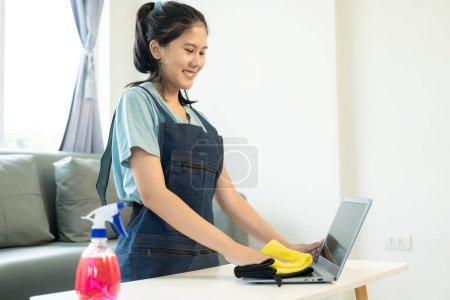 Téléchargez les photos : Femme asiatique en uniforme de ménage nettoie la maison et essuie la table avec un chiffon en microfibre dans le salon et vadrouille le sol avec un balai pour un intérieur propre et rangé. - en image libre de droit