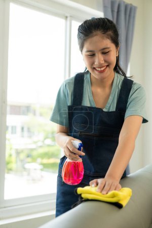 Téléchargez les photos : Femme asiatique en uniforme de ménage nettoie la maison et essuie la table avec un chiffon en microfibre dans le salon et vadrouille le sol avec un balai pour un intérieur propre et rangé. - en image libre de droit