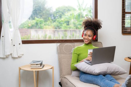 Photo for Young African American girl wearing headphones, smartphone, video call on laptop, looking at computer screen, watching webinar or doing video chat by webcam, online work concept. - Royalty Free Image