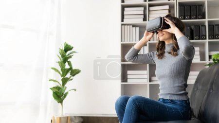 Téléchargez les photos : Joyeux jeune femme surprise dans le casque VR, sentir l'air. Femme caucasienne souriante portant des lunettes vr, dispositif de technologie de réalité virtuelle, regarder la vidéo de simulation 3D tout en étant assis sur le canapé à la maison. - en image libre de droit