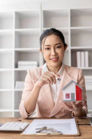 Téléchargez les photos : Les modèles de maisons près des représentantes asiatiques offrent des prix et des conditions d'achat ou de location sur des bureaux dans de nouveaux endroits. Signez le contrat - en image libre de droit