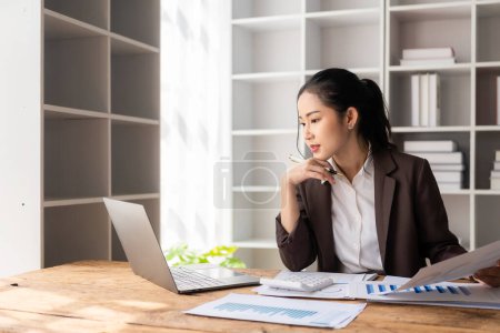 Foto de Hermosa mujer de negocios asiática analizando gráficos utilizando la calculadora en el ordenador portátil en la oficina con documentos sobre la mesa - Imagen libre de derechos