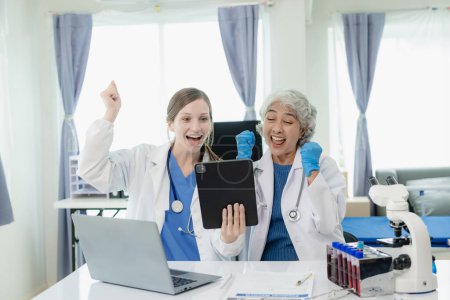 Photo for Successful Asian female doctor celebrates victory over successful treatment of patient in hospital, sitting at desk, arms raised with cheerful smile, research concept. - Royalty Free Image