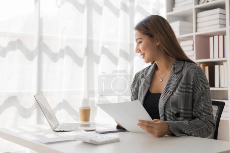 Photo for Asian female employee working using laptop computer at desk calculating expenses, financial reports on graph data sheet in office - Royalty Free Image