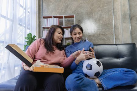 Foto de Jóvenes asiáticos sentados en el sofá viendo y animando el fútbol o el partido de fútbol en la televisión juntos en casa y celebrando la victoria. - Imagen libre de derechos