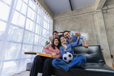 Photo for Young Asian men sitting on the sofa watching and cheering on football or football match on TV together at home and celebrating the victory. - Royalty Free Image