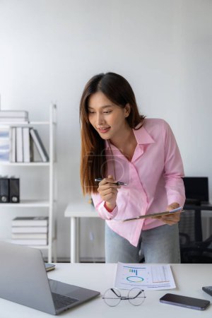 Photo for Young Asian woman sitting at desk with data and graph paper using laptop, analyzing accounts, planning strategies on business improvement in office. - Royalty Free Image