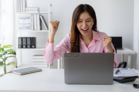 Téléchargez les photos : Heureuse femme asiatique célèbre le succès en levant les bras devant l'ordinateur portable. en serrant les poings Freelance, commencer à penser aux finances - en image libre de droit