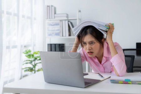 Photo for Young Asian female worker working with stress pretends to scratch her head because she is very tired working with laptop and documents on the table. - Royalty Free Image
