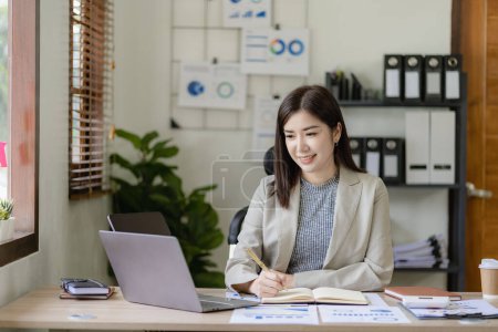 Téléchargez les photos : Ambitieuse fille asiatique travaillant à la maison En regardant l'écran d'ordinateur portable et les documents financiers, la femme vérifie les e-mails ou fait de la recherche tout en travaillant à distance. - en image libre de droit