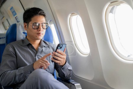 Foto de Un joven hombre de negocios asiático se sienta en un asiento cerca de la ventana hablando en su teléfono inteligente, la gestión de su negocio con su socio durante un vuelo para su viaje de negocios. - Imagen libre de derechos