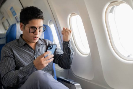 Photo for A young Asian businessman sits in a seat near the window talking on his smartphone, managing his business with his partner during a flight for his business trip. - Royalty Free Image