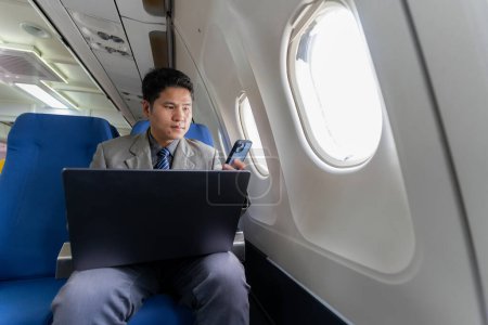 Foto de Un joven hombre de negocios asiático se sienta en un asiento cerca de la ventana hablando en su teléfono inteligente, la gestión de su negocio con su socio durante un vuelo para su viaje de negocios. - Imagen libre de derechos
