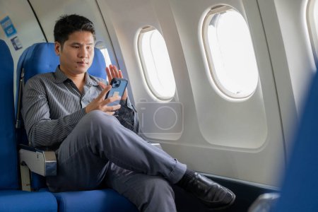 Téléchargez les photos : Un jeune homme d'affaires asiatique assis dans un siège près de la fenêtre parle sur son smartphone, gère son entreprise avec son partenaire lors d'un vol pour son voyage d'affaires. - en image libre de droit