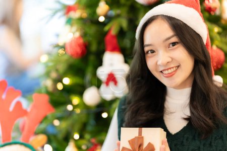 Téléchargez les photos : Jeune femme asiatique dans le chapeau de Père Noël souriant et tenant des cadeaux de Noël sourire heureux Tout en décorant le sapin de Noël à la maison, l'idée de célébrer le jour de l'An - en image libre de droit