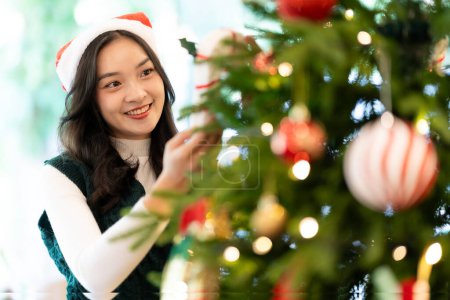 Téléchargez les photos : Jeune femme asiatique dans le chapeau de Père Noël souriant et tenant des cadeaux de Noël sourire heureux Tout en décorant le sapin de Noël à la maison, l'idée de célébrer le jour de l'An - en image libre de droit