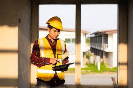 Foto de Joven ingeniero civil asiático o supervisor de construcción que usa un casco mira hacia otro lado y sonríe mientras inspecciona un sitio de construcción. - Imagen libre de derechos