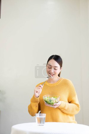 Foto de Sonriendo hermosa mujer joven le gusta comer ensalada de verduras frescas saludables para el desayuno. Dieta, desintoxicación, dieta, pérdida de peso, navegar por Internet en la sala de estar en casa. - Imagen libre de derechos