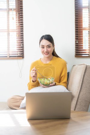 Photo for Smiling beautiful young woman likes to eat healthy fresh vegetable salad for breakfast. Dieting, detox, diet, weight loss, surfing the internet in the living room at home. - Royalty Free Image