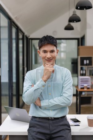 Photo for Young Asian businessman in his office and smiling with crossed arms. Young Asian businessman, successful professional, millennial, leader standing with arms crossed in front of computer screen in corporate office. - Royalty Free Image