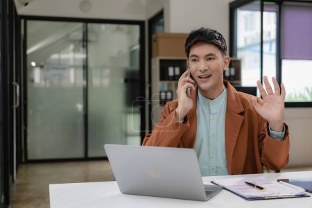 Téléchargez les photos : Jeune homme d'affaires asiatique en costume décontracté utilise un ordinateur portable et parle au téléphone pour communiquer avec les clients. réunion en ligne d'affaires en ligne - en image libre de droit