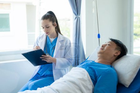 Téléchargez les photos : Un patient asiatique de sexe masculin se trouve dans un lit d'hôpital et est soigneusement soigné par un médecin. Médecin donnant des conseils au patient masculin travaillant sur le diagnostic de la maladie de santé - en image libre de droit