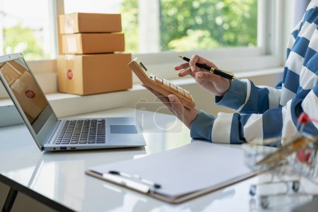 Photo for Asian woman working from home SME entrepreneurs use smartphones and laptops for commercial monitoring. Online marketing packed in a parcel box. Close-up image - Royalty Free Image