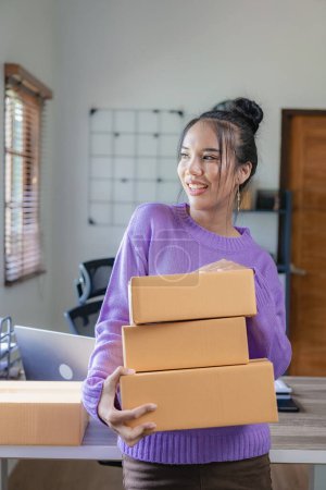 Photo for A purple-clad independent Asian woman SME uses a laptop and boxes to receive and review online orders to prepare packages to sell to customers. vertical picture - Royalty Free Image