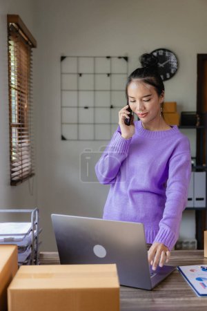 Téléchargez les photos : Une PME asiatique indépendante revêtue de violet utilise un ordinateur portable et des boîtes pour recevoir et examiner les commandes en ligne afin de préparer des colis à vendre aux clients. image verticale - en image libre de droit