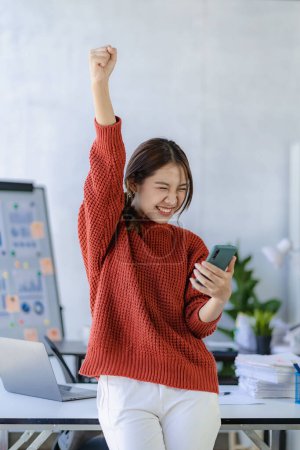 Téléchargez les photos : Employée asiatique excitée regardant l'écran du téléphone et heureux de recevoir de bonnes nouvelles de message dans l'application de chat sur téléphone mobile, concept d'entreprise. Image verticale - en image libre de droit