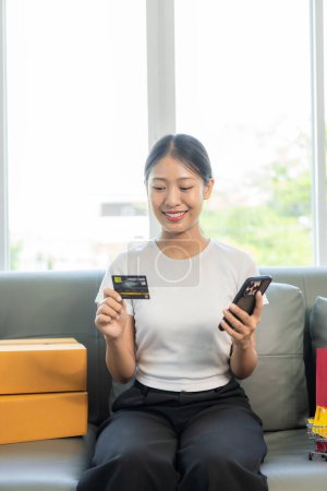 Téléchargez les photos : Femme asiatique joyeuse utilisant smartphone et ordinateur portable, achats en ligne, tenant la carte de crédit, payer, assis sur le canapé à la maison, concept de paiement via l'application bancaire en ligne. - en image libre de droit
