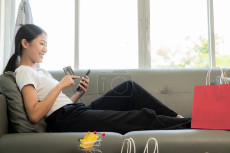 Téléchargez les photos : Femme asiatique joyeuse utilisant smartphone et ordinateur portable, achats en ligne, tenant la carte de crédit, payer, assis sur le canapé à la maison, concept de paiement via l'application bancaire en ligne. - en image libre de droit