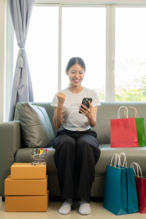 Photo for Cheerful Asian woman using smartphone and laptop, online shopping, holding credit card, paying, sitting on sofa at home, payment concept through internet banking application. - Royalty Free Image