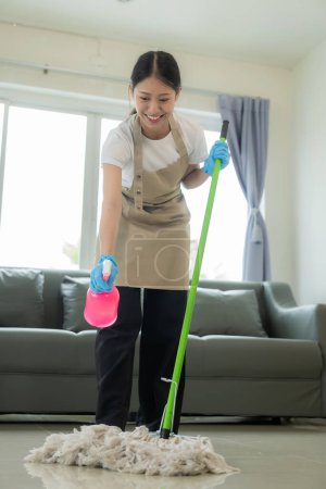 Téléchargez les photos : Belle femme au foyer asiatique faisant des travaux ménagers et le nettoyage de la maison avec un tablier, en utilisant un aspirateur, une serpillière, un chiffon pour nettoyer le sol dans le salon. Bonne femme au foyer faisant des travaux ménagers - en image libre de droit