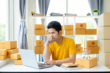 Téléchargez les photos : Un jeune homme asiatique propriétaire d'une PME vend des produits en ligne et prépare des boîtes d'emballage de produits pour envoyer des commandes en ligne et avoir des clients à la maison qui achètent en ligne prêt pour la livraison. - en image libre de droit
