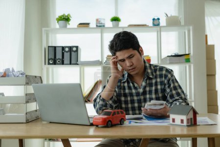 Photo for A young Asian man is stressed about bills, car payments, house payments, and holds his head in his hands as he looks at the bills. Confused about financial information or worried about spending money - Royalty Free Image