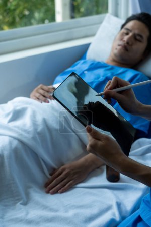 Photo for Young Asian male patient lying in bed while checking his pulse. Consult and explain with the nurse who takes notes and assists in the hospital in the recovery room in the hospital department. - Royalty Free Image