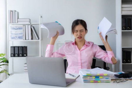 Photo for Young Asian woman puts her hands on her head feeling tired, irritated and stressed from hard work at the office and dissatisfied with her job while looking at report documents. - Royalty Free Image