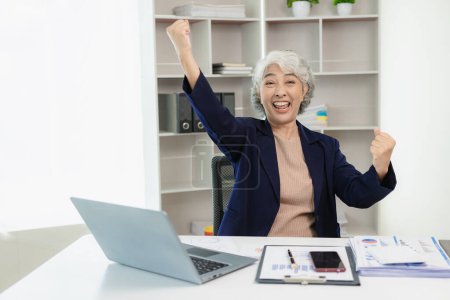 Téléchargez les photos : Très excité asiatique senior femme d'affaires heureux en utilisant un ordinateur portable pour recevoir de bonnes nouvelles. Entrepreneur excité obtient une promotion ou complète un projet. Heureux de leur propre travail - en image libre de droit