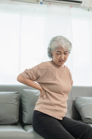Foto de Una anciana tiene dolor de espalda después de despertarse mientras está sentada en el sofá de su casa. Una mujer asiática de edad avanzada tiene dolor en la cintura o las nalgas después de caer. - Imagen libre de derechos