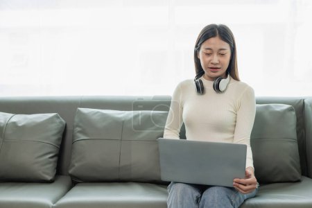 Foto de Mujer asiática joven usando el ordenador portátil y sonriendo mientras se sienta en el sofá de trabajo desde casa Trabajando de forma remota, el nuevo concepto normal y abrazando Internet - Imagen libre de derechos