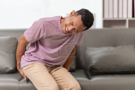 Junger Mann mit Hämorrhoiden sitzt auf dem Sofa und berührt ihr Gesäß wegen Bauchschmerzen und Hämorrhoiden Gesundheitskonzept.