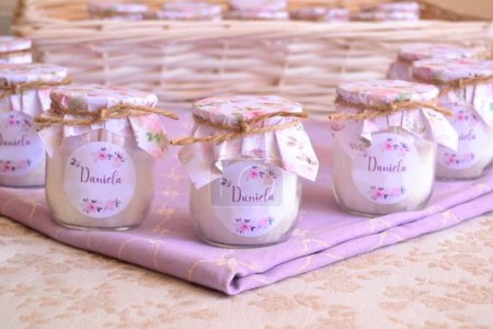 Baptism baby girl shower favors soy candles in glass yogurt jar with floral pattern wrapping paper, jute ribbon and pink custom label text baby name,white wicker and purple background, original diy handmade party souvenir