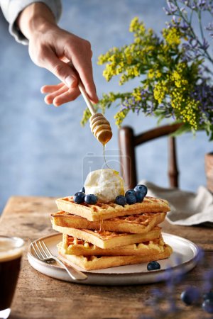 Photo for Waffles topped with blueberries, ice cream and honey standing on a rustic brown table. Blue background. Hand drizlling honey. - Royalty Free Image