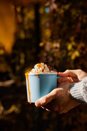 Photo for Woman's hand holding a blue cup of hot chocolate with whipped cream and caramel in the autumn forest. - Royalty Free Image
