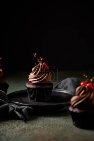 Téléchargez les photos : Magnifique cupcake au chocolat avec glaçage au chocolat garni de groseilles rouges et d'une branche. Style rustique. Placé sur une plaque et une serviette en céramique noire sur fond de pierre verte. - en image libre de droit