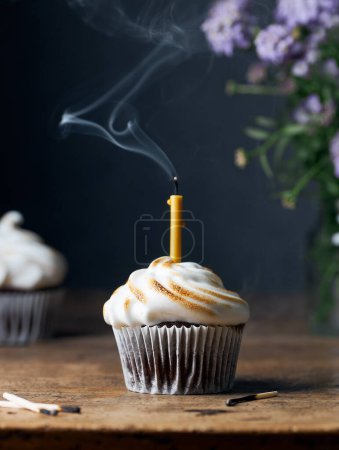 Photo for Chocolate cupcakes with meringue frosting on a rustic old wooden table. One cupcake has blown out candle. Candle still has smoke. Black background. Purple flowers in the background. Moody vertical still life with space for copy. - Royalty Free Image